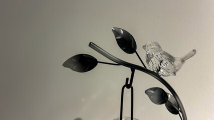 Elegant decorative ornament featuring a transparent glass bird perched on a black metal branch with leaves. The design creates a minimalist and artistic contrast against a neutral background, symbol
