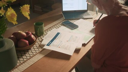 Over the shoulder view of senior woman looking at gas bill and writing meter reading down when calculating monthly expenses with laptop and smartphone at kitchen table