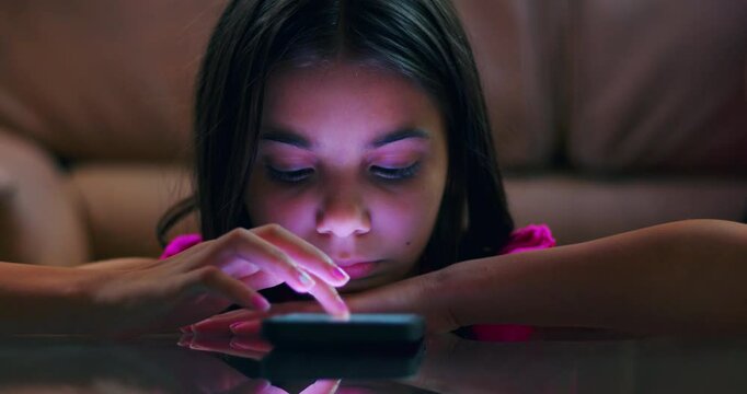 Young girl resting her head on arms while using smartphone on glass table, focused and engaged with screen in dim indoor setting, glowing light on face
