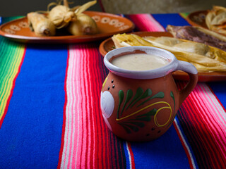 Clay cup with atole over colorful serape. Tamales in the background. Mexican food
