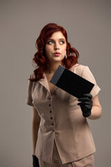 Naklejka premium Confident woman with red hair, holding a black envelope, wearing beige outfit and gloves, looking away, modern fashion concept