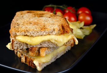 A sandwich with grilled cheese on the black plate. cherry tomato and crisps on the side. The sandwich is cut in half and the cherry tomato are on the side