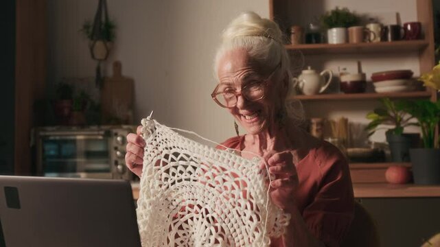 Tilt up rack focus shot of joyful Caucasian senior woman saying hello to her friend and showing them crochet piece when video calling in front of laptop at kitchen table
