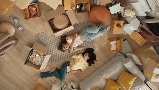 Lesbian lover looking at phone while laughing together and lying down at living room floor with cardboard box scattering. Happy couple checking at smart phone while spending time together. Myrmidon.