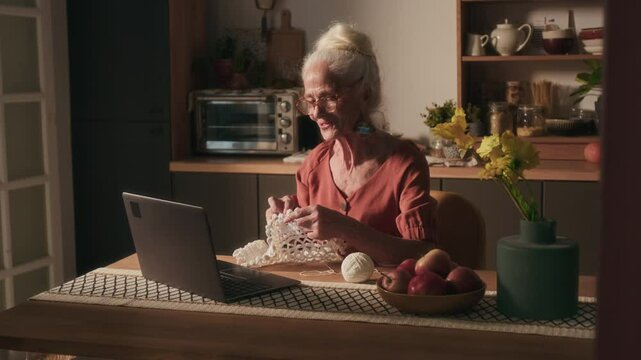 Cheerful elderly lady waving at laptop camera and showing her crochet piece during video call with friend when sitting at wooden table with ball of yarn in kitchen