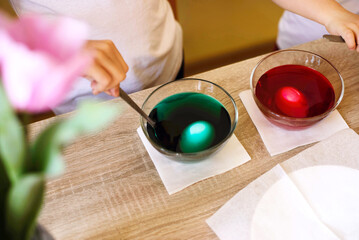 Children dying Easter eggs in vibrant red and green colors. Children's hands mixing paint in glass bowls on table. Festive holiday tradition, celebrating joy of Easter preparations and upcoming spring