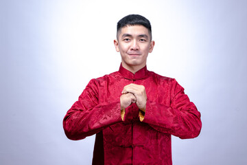 A smiling young man in a red traditional Chinese Tang suit performs a Gong Xi greeting gesture,...