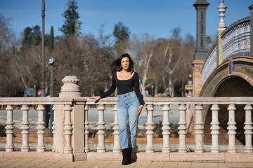 Fototapeta premium Young, beautiful, brunette woman, wearing black long sleeve t-shirt and jeans, with her hair in the wind, posing leaning on a railing in the square of Spain in Seville. Concept beauty, femininity.