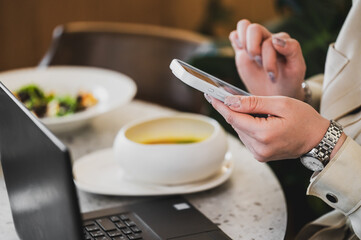 A person is using a smartphone while sitting at a table with a laptop and a bowl of soup, reflecting a modern work-life balance and casual dining atmosphere.