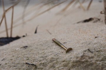 A detailed macro image of a golden screw resting in beach sand. The screw's metallic gleam stands out against the natural, fine texture of the sand, offering an intriguing contrast between man-made
