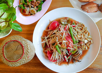 Top view papaya salad (Som Tam) Popular traditional Thai food with spicy flavors in white plate and served with sticky rice and fresh vegetables. on wooden table.