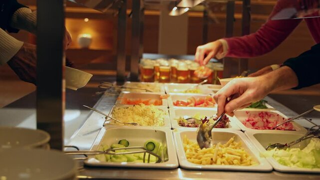 People helping themselves at the colorful salad bar with a healthy selection of fresh vegetables, glasses with colorful desert in the background