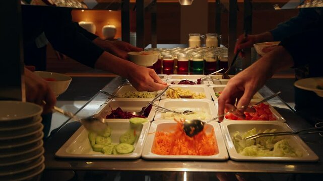 People helping themselves at the colorful salad bar with a healthy selection of fresh vegetables, glasses with colorful desert in the background
