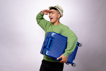 An Asian man in a green sweater and a fedora hat holds a blue suitcase while looking into the distance with an excited expression. He appears ready for an adventure, posing against a white background