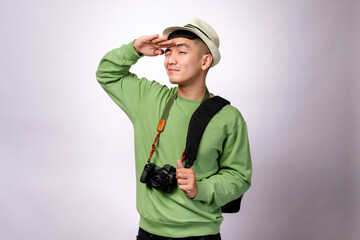 An Asian man in a green sweater, beige hat, and black shoulder bag saluting playfully while looking into the distance. He has a camera around his neck and is smiling. The background is white