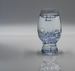 A clear glass filled with water and ice cubes sits on a smooth, reflective surface. The light highlights the clarity of the liquid and the ice, creating a refreshing appearance.