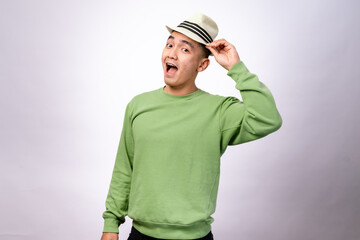 An Asian man in a green sweater is playfully holding a fedora hat above his head while smiling. He looks cheerful and stylish against a plain white background