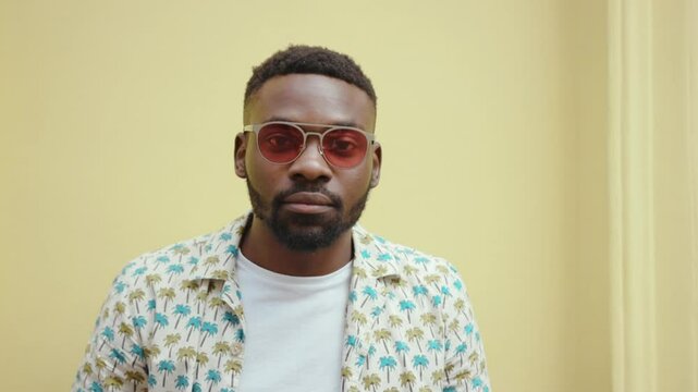 Good-looking young African man wearing stylish clothes putting sunglasses with red lenses. Focused guy looking directly at camera. Yellow wall.