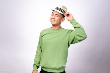 An Asian man in a green sweater is playfully holding a fedora hat above his head while smiling. He looks cheerful and stylish against a plain white background