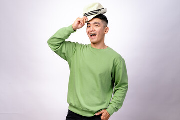 An Asian man in a green sweater is playfully holding a fedora hat above his head while smiling. He looks cheerful and stylish against a plain white background