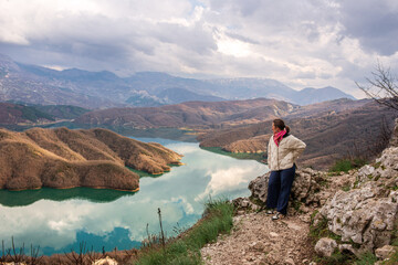 A woman in a white jacket with a pink scarf stands on the edge of a cliff, gazing at a stunning...