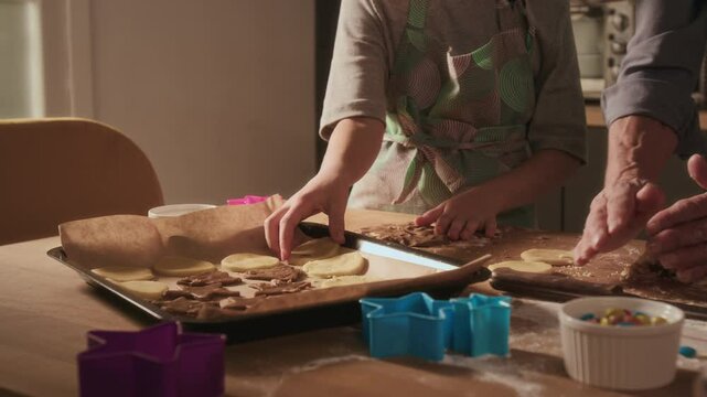 Midsection view of unrecognisable girl wearing kitchen apron placing dough circles on parchment paper while her grandmother collecting leftover dough