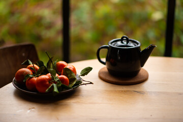 Fresh clementines with green leaves in a plate on a wooden table. tangerines and a teapot with tea in a rustic style