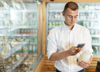 Attentive young man purchaser scanning QR code on bottle of milk standing in supermarket with...