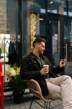 Vertical portrait of happy young man sitting at outdoor cafe chair, holding takeaway coffee cup and browsing smartphone, enjoying relaxing break in vibrant urban atmosphere. Concept of city lifestyle.