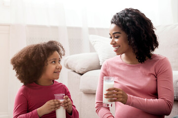 Healthy black mom and cute little daughter sitting with glass of milk on floor in living room, spending time together at home, free space