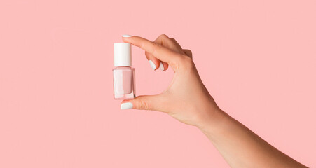 Unrecognizable young girl holding bottle of nail polish on pink background, close up with copy space. Panorama