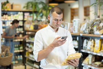 Attentive young man purchaser scanning QR code of pasta in big supermarket with large assortment