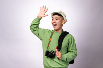 An Asian man in a green sweater, fedora hat, and camera around his neck waves happily while carrying a black shoulder bag. He looks cheerful and excited, standing against a plain white background