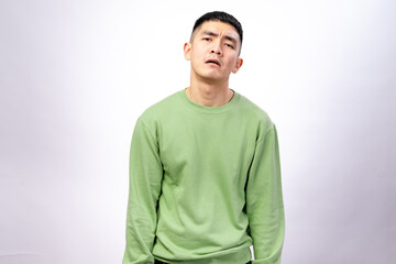 An Asian man in a green sweatshirt stands against a white background, slouching with a tired,...