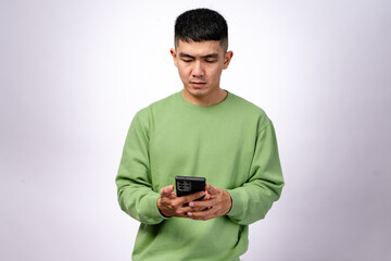 An Asian man in a green sweatshirt is focused on his smartphone, holding it with both hands while looking at the screen. His expression is neutral. The background is white