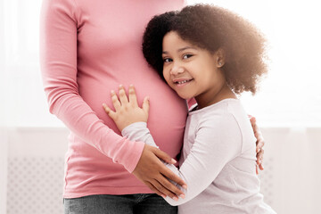 Fototapeta premium Close up of little black daughter hugging her pregnant mother's tummy, standing next to window at home