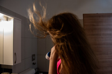 Fototapeta premium Young woman in a pink top uses a hairdryer, flipping her hair back as warm air flows through it. The dimly lit bathroom creates a dramatic and stylish effect.