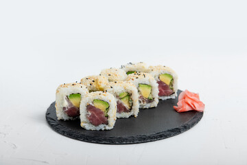 Close-up of sushi California rolls filled with tuna, avocado, and sesame seeds served on a black slate plate, accompanied by pickled ginger on isolated white background