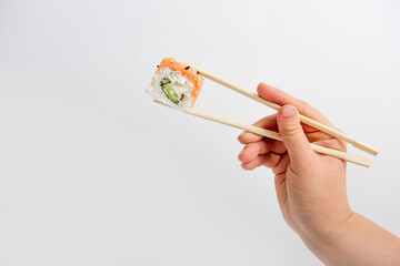 Hand holding a fresh sushi Philadelphia roll with chopsticks against a white background. The roll contains rice, salmon, avocado, and cream cheese, topped with sesame seeds.