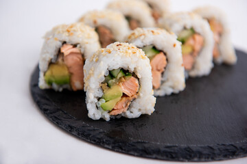 Fresh sushi rolls with salmon, avocado, and cucumber, topped with sesame seeds. Served on a black slate plate for an elegant presentation.