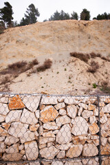 Close-up of a gabion wall made of irregular stones held by a metal mesh, set against a rugged hillside with sparse vegetation and trees in the background.