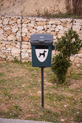 Green dog waste bin with a black lid and dog symbol, placed in a grassy area near a stone wall and bush. Designed for pet waste disposal in public spaces.