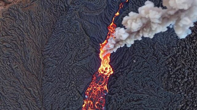 A dramatic aerial shot of an active volcano shows molten lava streaming down its rugged slopes, surrounded by hardened black volcanic rock, with thick smoke rising into the sky