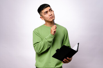 An Asian man in a green sweater is thinking while holding a pen near his chin and a black notebook...