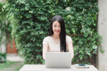 Girl working at desk outdoor: A beautiful young woman works on a laptop at a table, surrounded by a lush, green garden. Her eyes are fixed on the computer with joy and is smiling. 