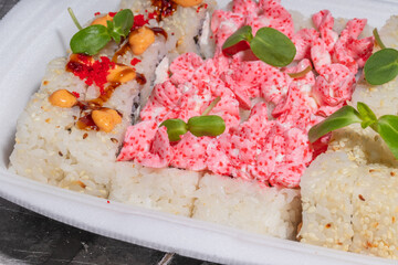 A detailed close-up of sushi rolls with pink creamy seafood topping, spicy sauce, and green garnish, served in a takeout box.