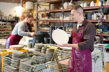 Positive skilled male artisan standing amidst racks with collection of crafted wares in sunlit...