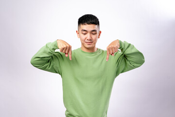 An Asian man in a green sweatshirt points downward with both index fingers, looking focused and slightly serious. He stands against a plain white background, conveying direction, instruction