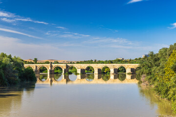 Fototapeta premium Roman bridge in Cordoba town in Spain. Ancient stone bridge spanning a calm river, reflecting the clear blue sky.
