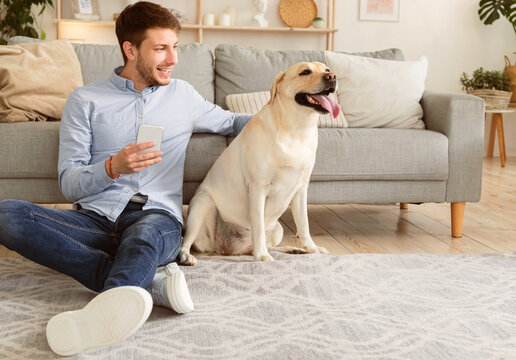 Spending Time At Home Concept. Handsome guy using smart phone sitting on the carpet and embracing his pet